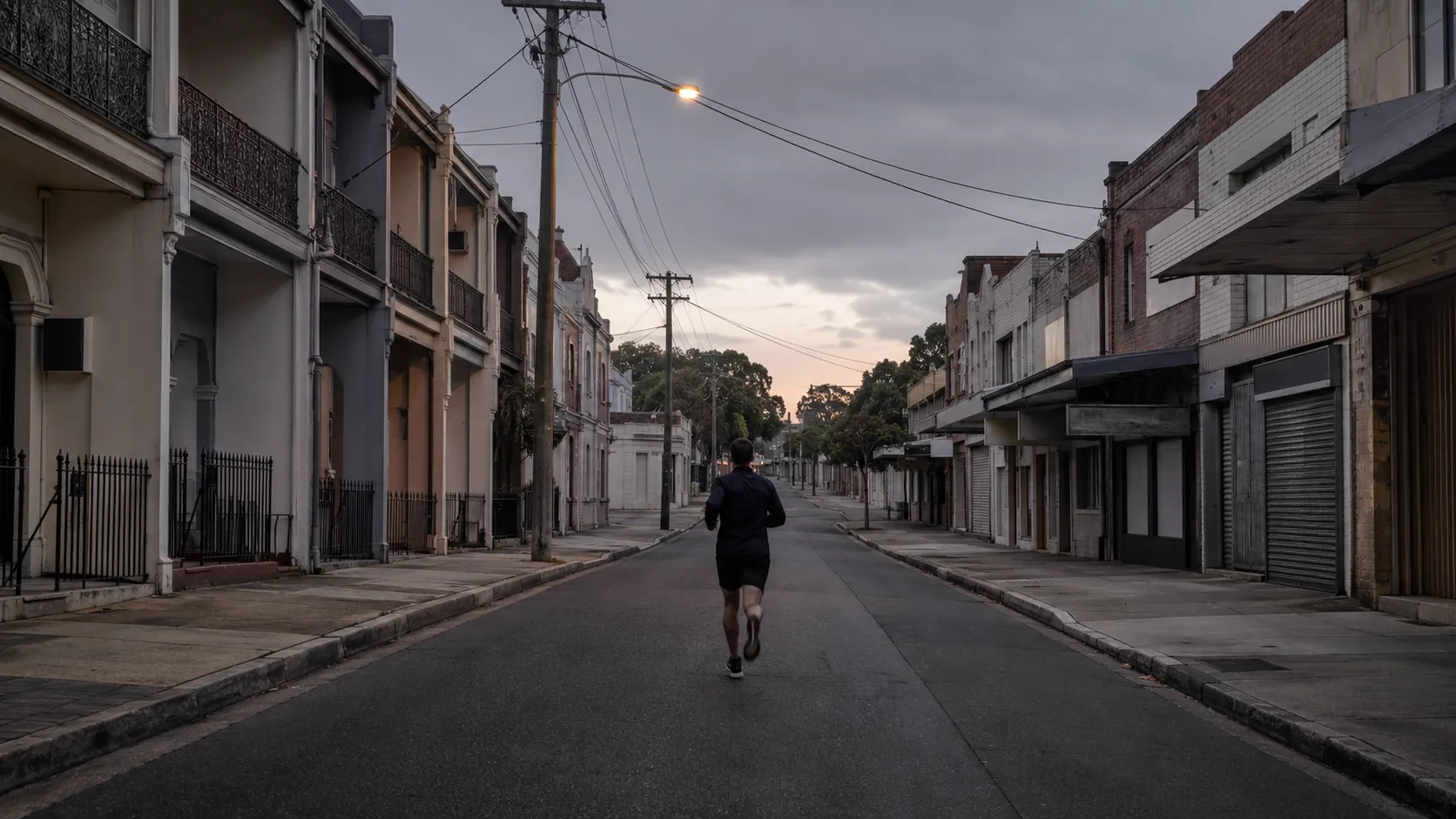 Michael Le running alone down an empty Inner West Sydney street at dawn during lockdown, terrace houses and shut shopfronts either side, one streetlight still on.