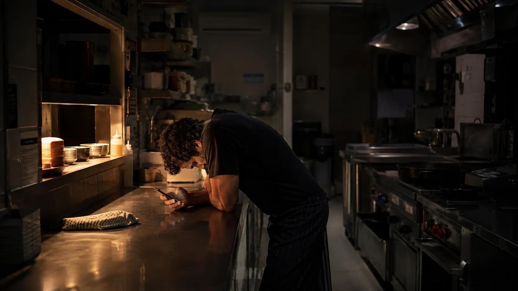 A restaurant owner alone in his kitchen after close, leaning on the bench with his head down and phone in hand, one pass light still on.
